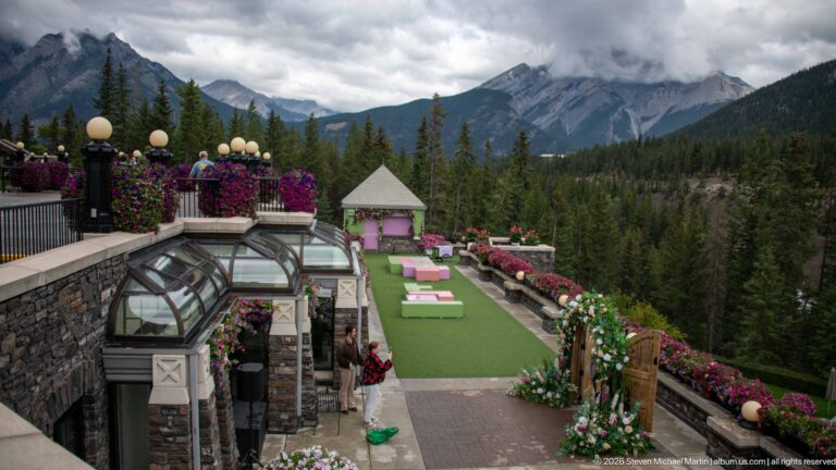 Tiered hotel terrace with green lawn, pink furnishings, flower-lined stone walls, glass roof, forest, and clouded mountain beyond.