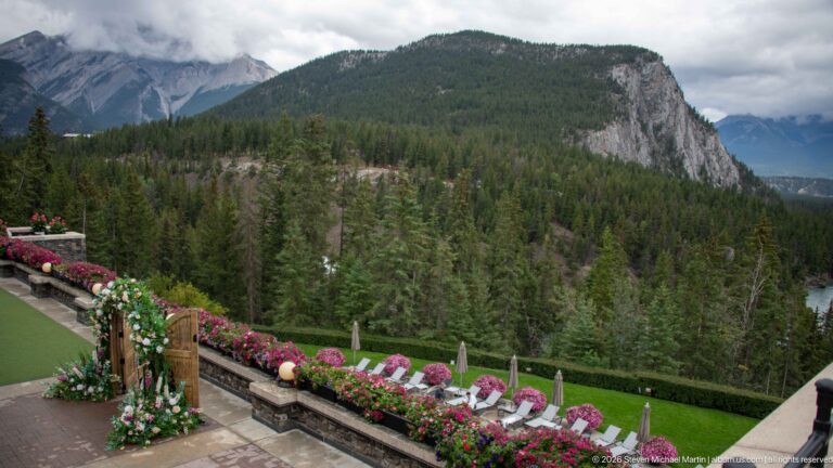 Stone hotel terrace with flower baskets and lounge chairs overlooking dense forest and a rocky mountain escarpment.