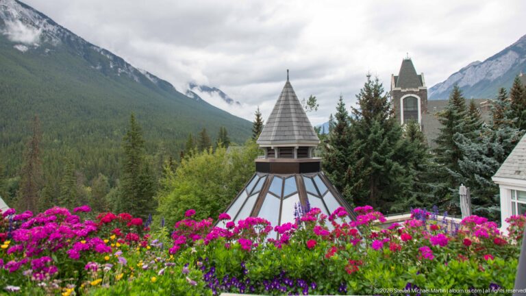 Small steep-roofed turret rising behind vivid flower beds, with trees, another tower, and cloudy mountains in the distance.