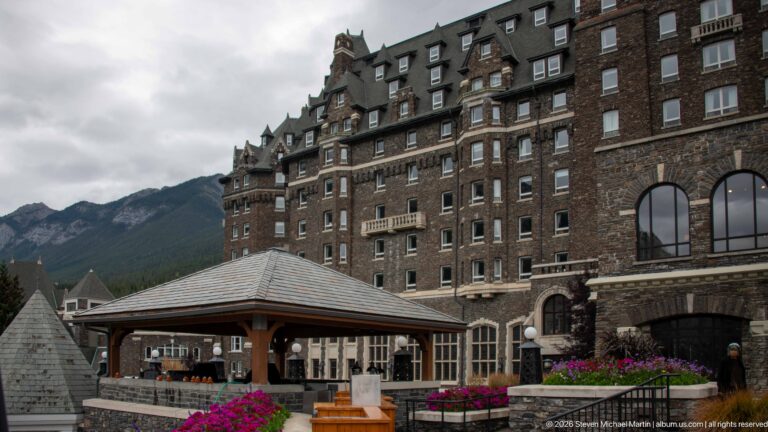 Timber-roofed terrace pavilion below the tall stone Banff Springs Hotel façade with windows, dormers, balconies, and flowers.