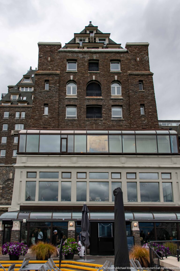 Banff Springs Hotel terrace-side wall with dark masonry, broad glazed enclosure, balconies, stepped roofs, umbrellas, and planters.