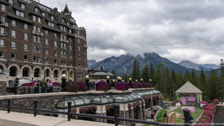 Tiered Banff Springs Hotel terraces with stone walls, glass roofs, flowers, lawn, guests, and mountains beyond.