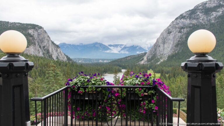 Flowered terrace railing between two globe lamps overlooking a forested valley, river, cliffs, and distant cloud-wrapped mountains.