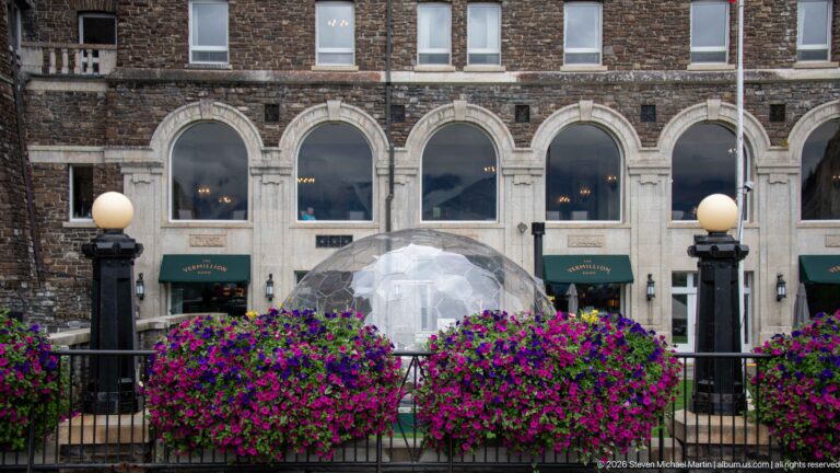 Transparent dining dome behind magenta flowers and black globe lamps, beneath large arched windows of Banff Springs Hotel.