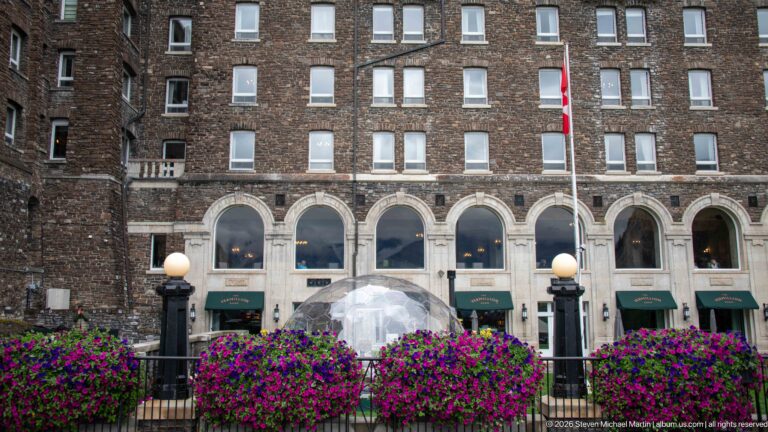 Dark masonry hotel façade with pale arched windows, green awnings, Canadian flag, globe lamps, flowers, and dining dome.