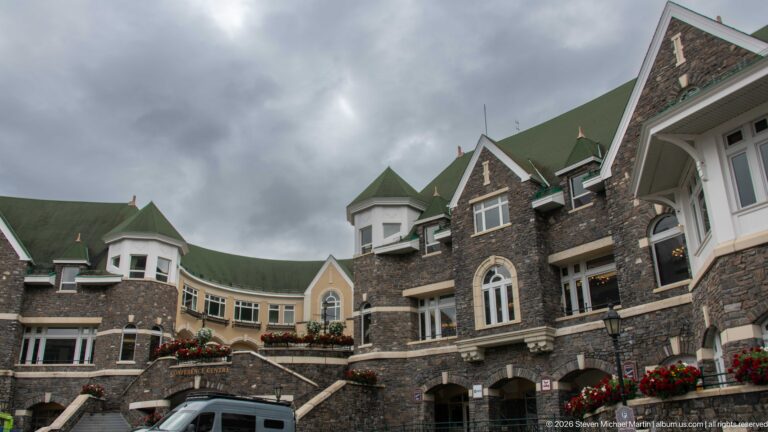 Hotel wing with dark stone walls, pale stucco sections, green gabled roofs, projecting bays, exterior stairs, and flower boxes.