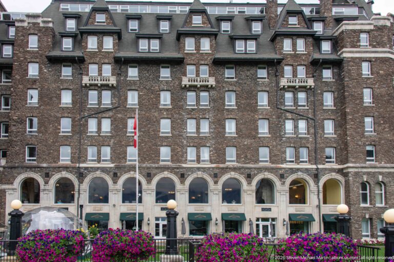 Banff Springs Hotel façade with rows of windows, pale stone arches, green awnings, hanging flowers, and a transparent dining dome below.