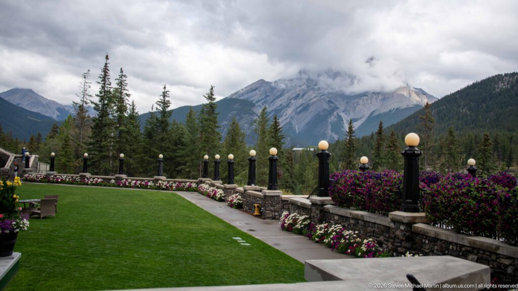 Formal green lawn and flower-lined stone terrace with globe lamps overlooking forested Rocky Mountain peaks under low clouds.