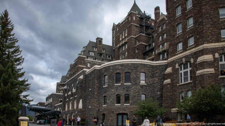 Ground-level view along Banff Springs Hotel stone wings with arched openings, towers, varied rooflines, and pedestrians below.