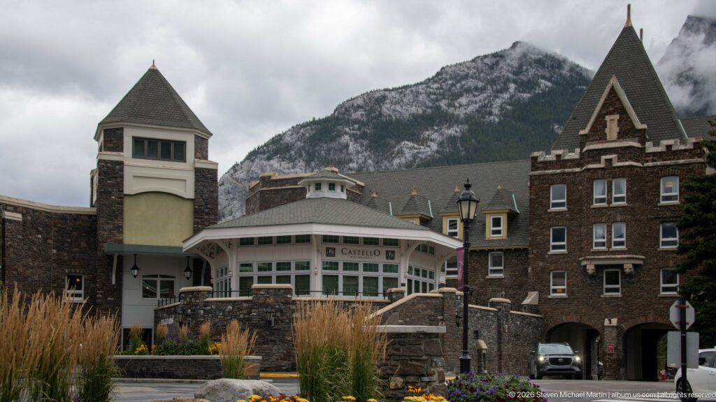 Castello Italiana entrance with curved windows, square turret, dark-stone hotel wing, flowers, and mountains behind.