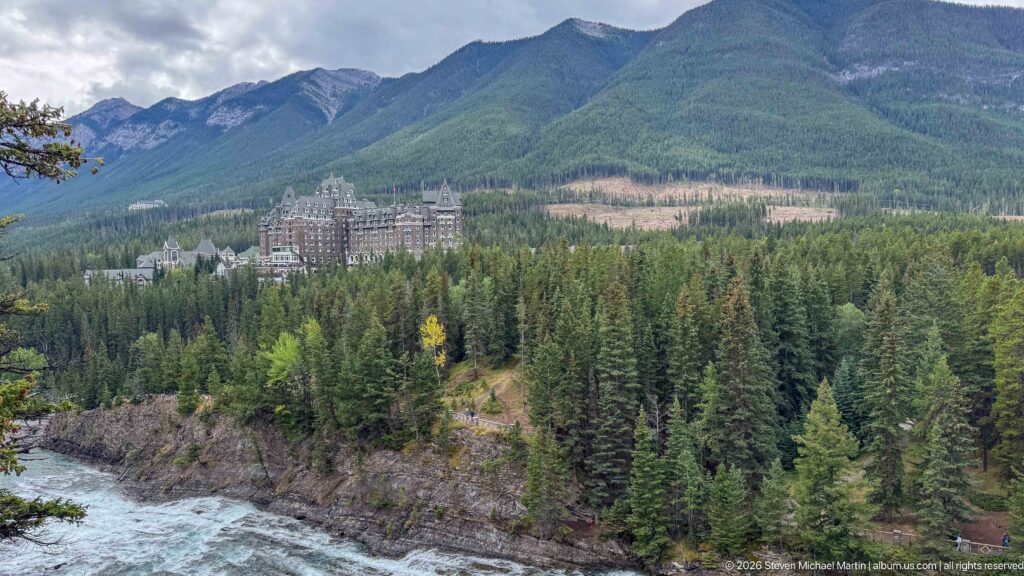 Distant Banff Springs Hotel across the Bow River gorge, surrounded by evergreen forest and backed by mountain slopes.