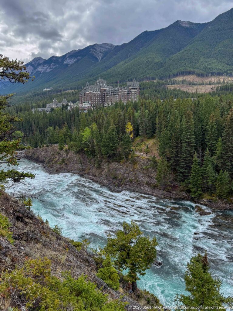 Wide Bow River bend with rocky banks and evergreen forest, Banff Springs Hotel small in the distance beneath mountains.