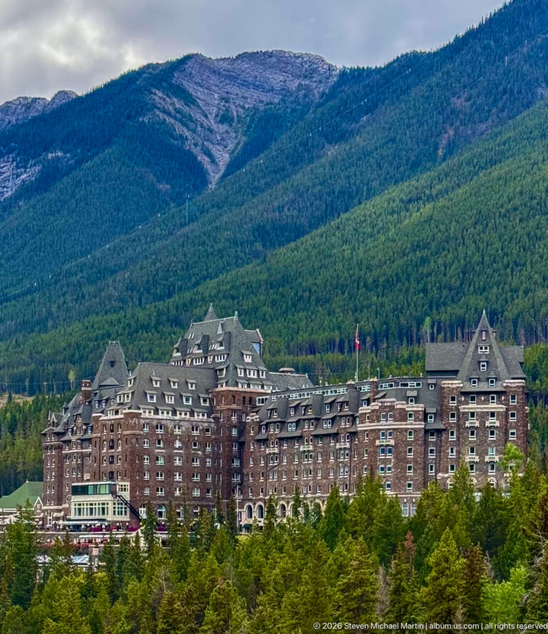 Broad view of Banff Springs Hotel above treetops, with long masonry wings and steep roofs backed by a forested mountain.