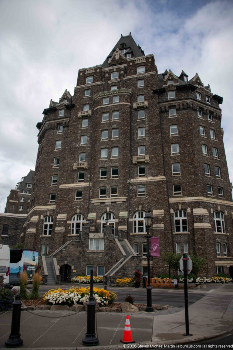 Low-angle view of a massive dark-stone hotel façade with rows of windows, steep roofs, steps, flower beds, vehicles, and bollards.