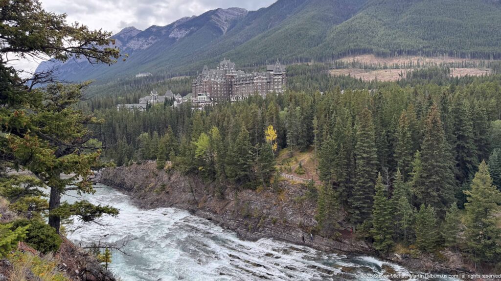 Bow River rapids in the foreground with Banff Springs Hotel across dense forest below green mountain slopes.