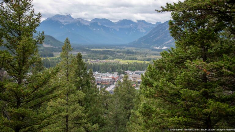 Forested overlook toward the Bow Valley and Banff townsite, with layered Rocky Mountain peaks beneath a cloudy sky.