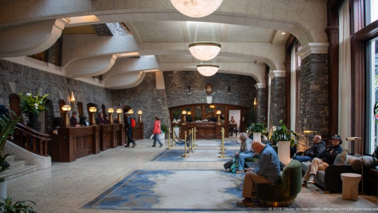 Hotel reception hall with front desks, seated guests, tall windows, blue rugs, stone walls, pendant lights, and an archway beyond.