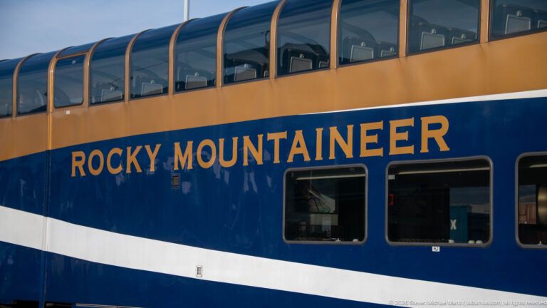 Blue, white, and gold Rocky Mountaineer bi-level passenger car with large upper panoramic windows and visible observation seats.