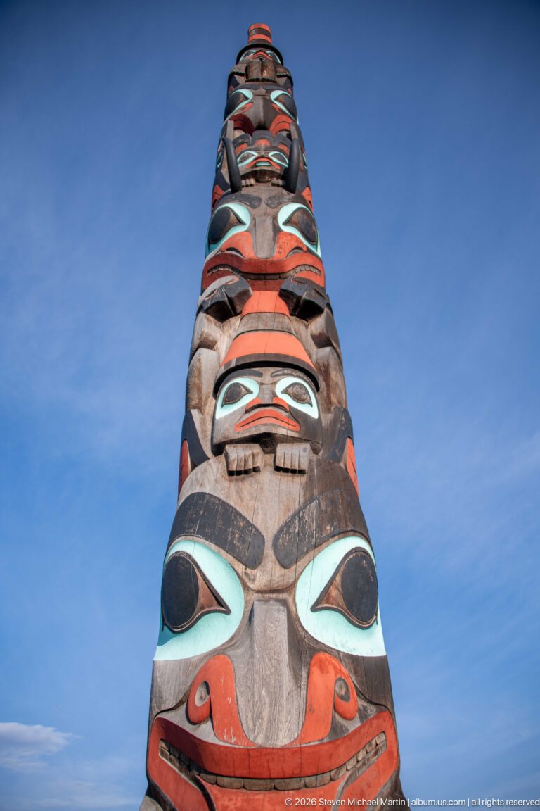 Tall carved and painted totem pole viewed steeply upward, with stacked figures receding into a blue sky.