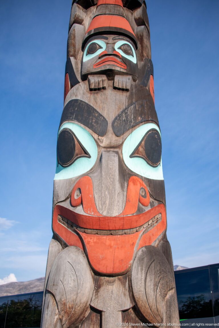 Low-angle close-up of a carved totem-pole figure painted in red, black, pale blue, and natural wood tones.