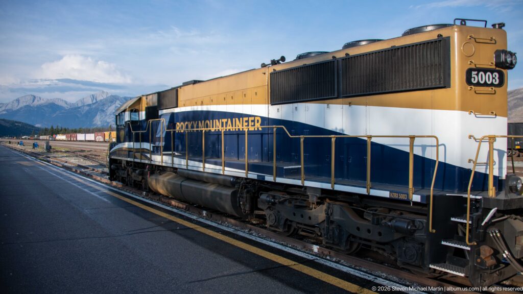 Blue, white, and gold Rocky Mountaineer locomotive 5003 beside a platform with rail yard and mountains behind.