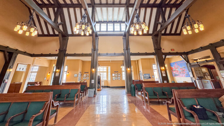Wide station interior with green bench rows, tall dark timber posts, exposed trusses, pendant lights, and a central aisle.
