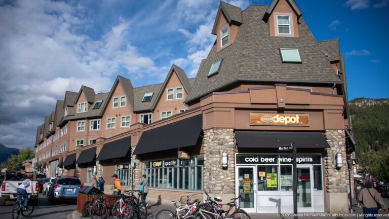 Cool Depot corner shop beneath a tall pointed gable, with repeating dormers, black awnings, bicycles, and parked cars.