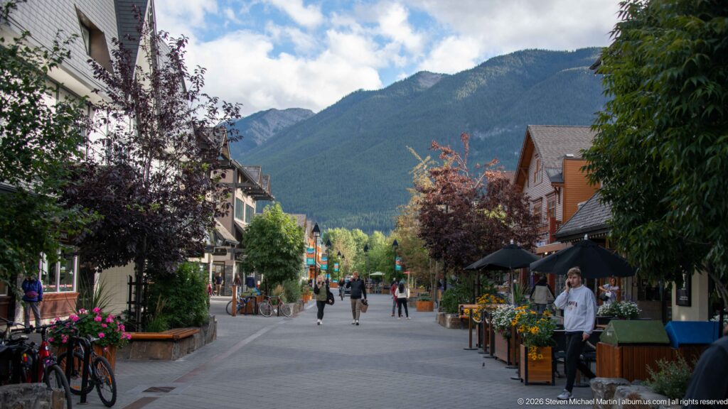 Walkers on a pedestrian corridor lined with bicycles, restaurant terraces, flowers, trees, and mountains ahead.