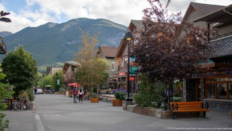 Pedestrian street lined with shops, benches, bicycles, trees, and café seating beneath a forested mountain.
