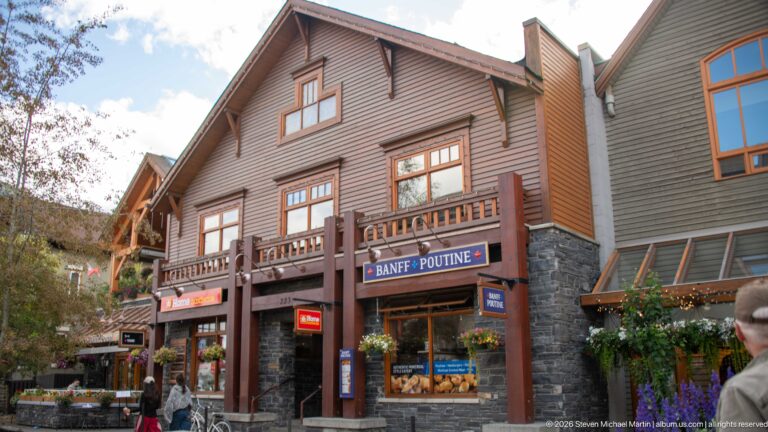 Steep-gabled brown commercial building with stone base, timber balconies, black awnings, and a Banff Poutine storefront.