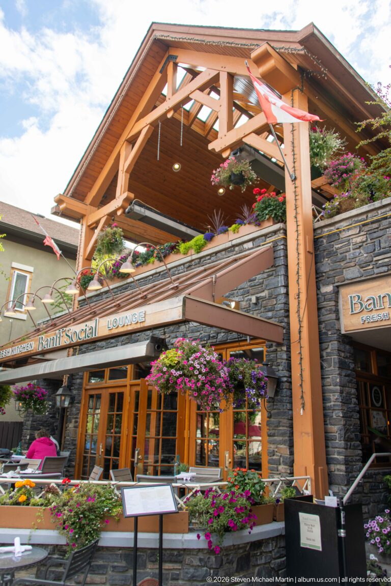 Timber-and-stone Banff Social restaurant façade with flower baskets, Canadian flag, terrace, and menu stands.