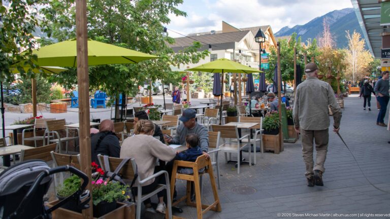 Outdoor café seating and pedestrians along a landscaped commercial street in Banff, with a mountain rising beyond the buildings.