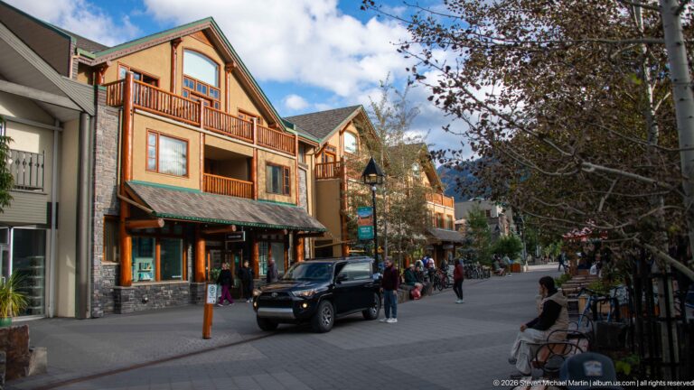 Black pickup moving through a pedestrian-filled Banff lane lined with timber balconies, shops, trees, and outdoor seating.