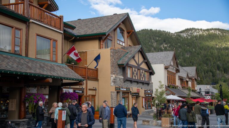 Pedestrians beside lodge-style Banff shops displaying Canadian and Alberta flags, with a forested mountain behind.
