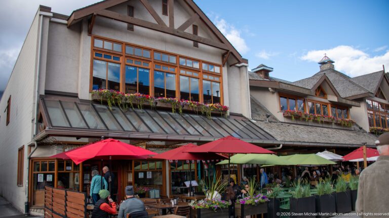 Restaurant terrace with red umbrellas beneath a gabled, half-timbered building with flower boxes and large windows.