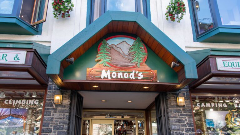 Monod’s entrance with carved mountain-and-tree sign, small gable, stone facing, and outdoor-gear displays.