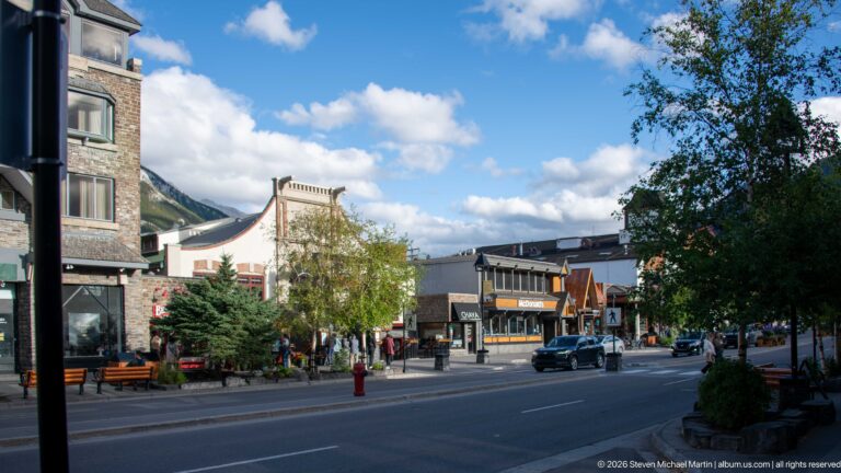 Wide Banff street lined with shops, trees, pedestrians, vehicles, and mountain slopes beneath blue sky.