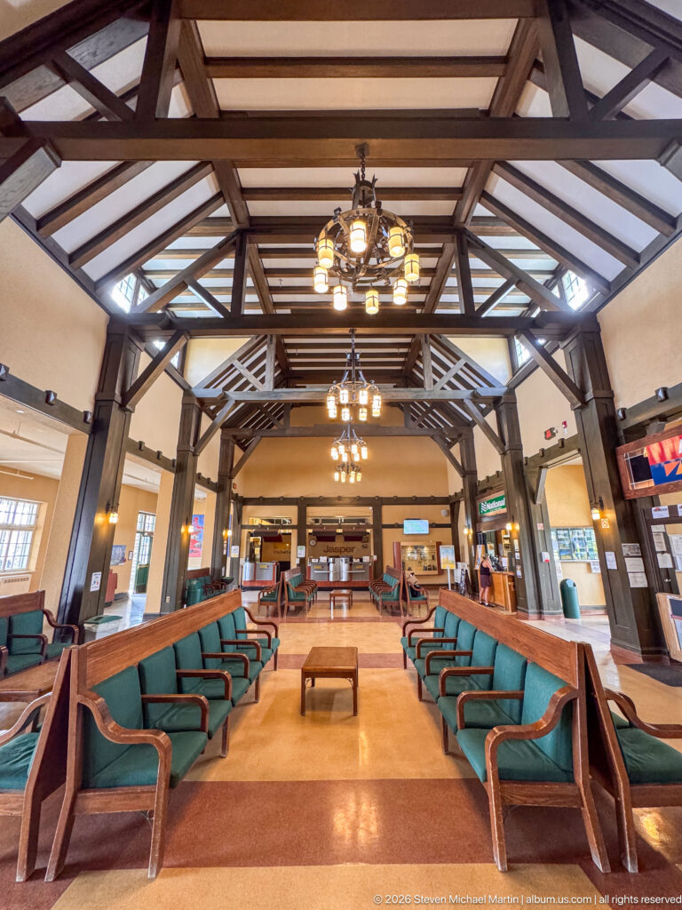 Symmetrical railway waiting room with exposed dark timber trusses, hanging lights, green benches, and service counters beyond.