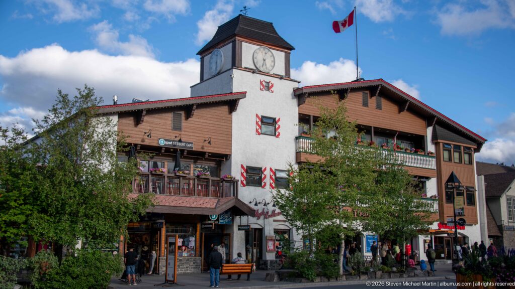 Large Banff commercial building with white clock tower, Canadian flag, balconies, shutters, shops, and pedestrians.