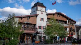 Large Banff commercial building with white clock tower, Canadian flag, balconies, shutters, shops, and pedestrians.