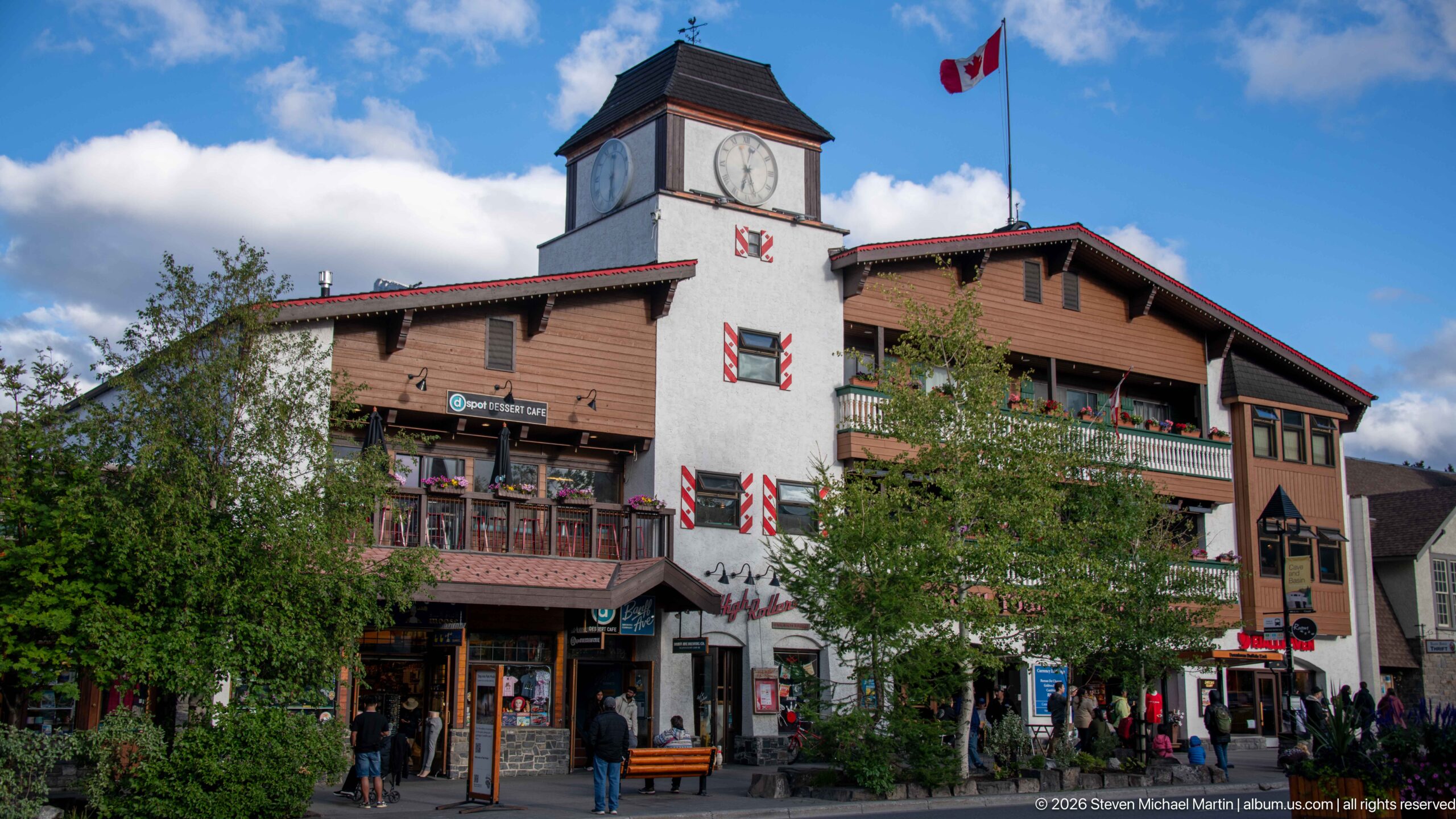 Large Banff commercial building with white clock tower, Canadian flag, balconies, shutters, shops, and pedestrians.