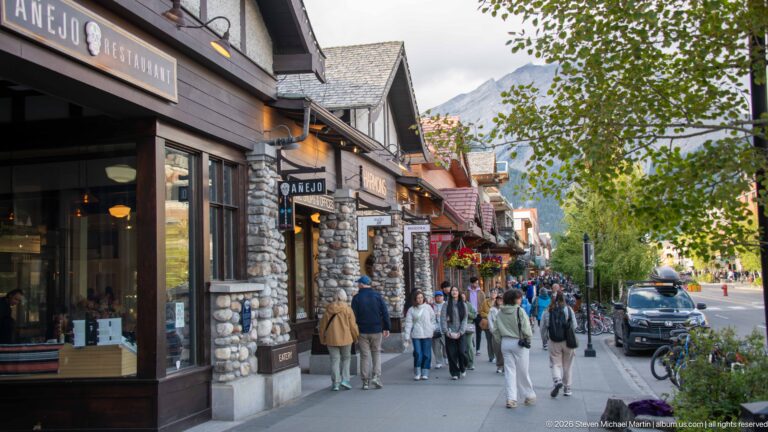 Busy Banff sidewalk beside stone-and-timber shops, hanging signs, flowers, bicycles, and pedestrians.