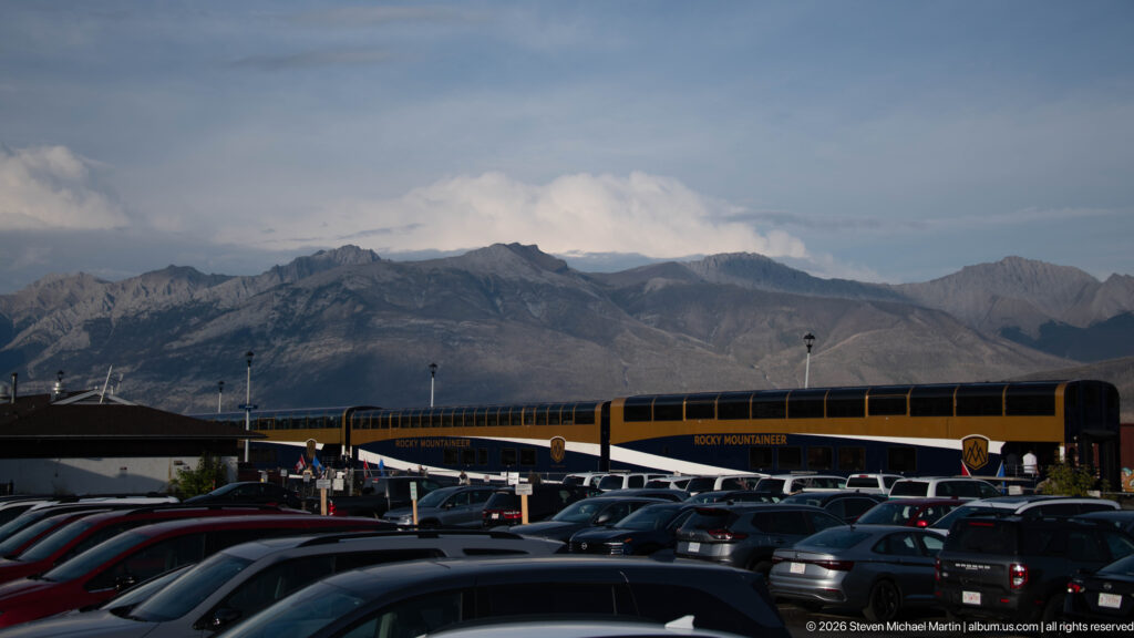 Long Rocky Mountaineer train beyond a parking area, with rugged Canadian Rockies peaks filling the background.