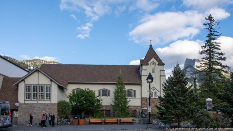 White stucco church with long gabled nave, square corner tower, multi-pane windows, and trees.