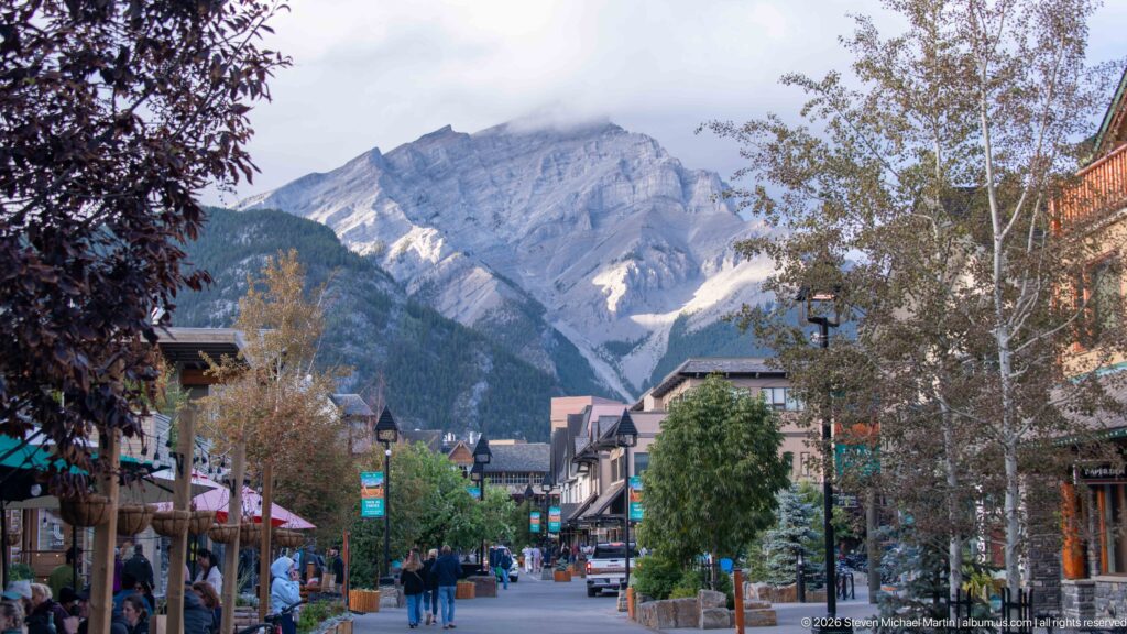 Pedestrians on a Banff commercial street with shops and trees below cloud-covered Cascade Mountain.