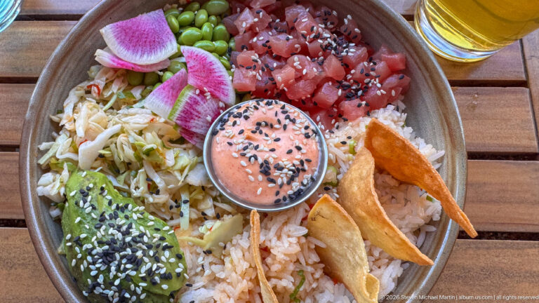 Overhead view of rice bowl with diced tuna, avocado, edamame, radish, vegetables, chips, and sauce.
