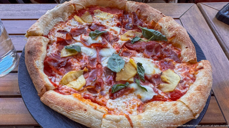 Round pizza topped with tomato, cheese, cured meat, artichokes, and basil on a dark serving plate.