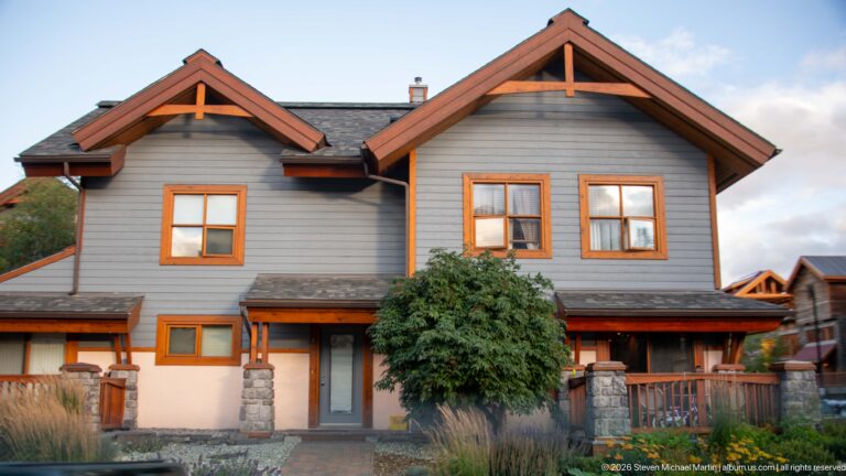Front of gray gabled house with orange windows, stone porch piers, central door, and timber braces.