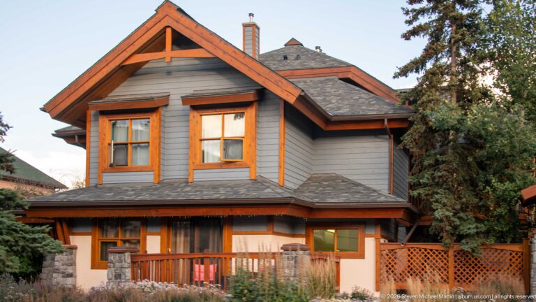 Gray two-story house with orange trim, gabled roofs, porch, stone base, and mature trees.