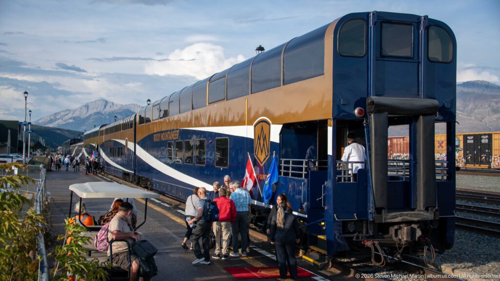 Passengers gather beside the open rear end of a Rocky Mountaineer car, with the train and mountains extending behind.